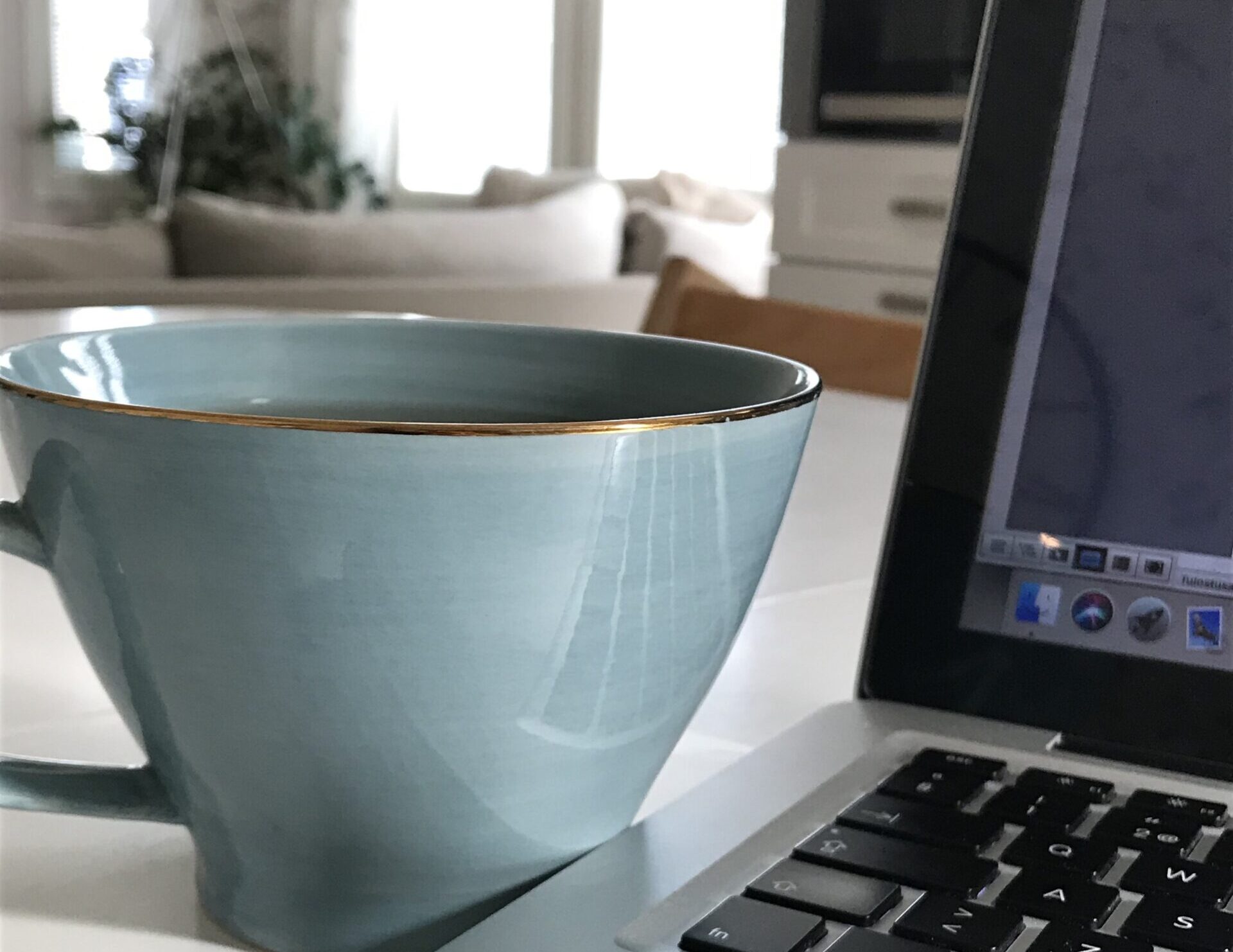 A pale blue cup with a gold rim on a white desk, with a laptop softly visible in the background.